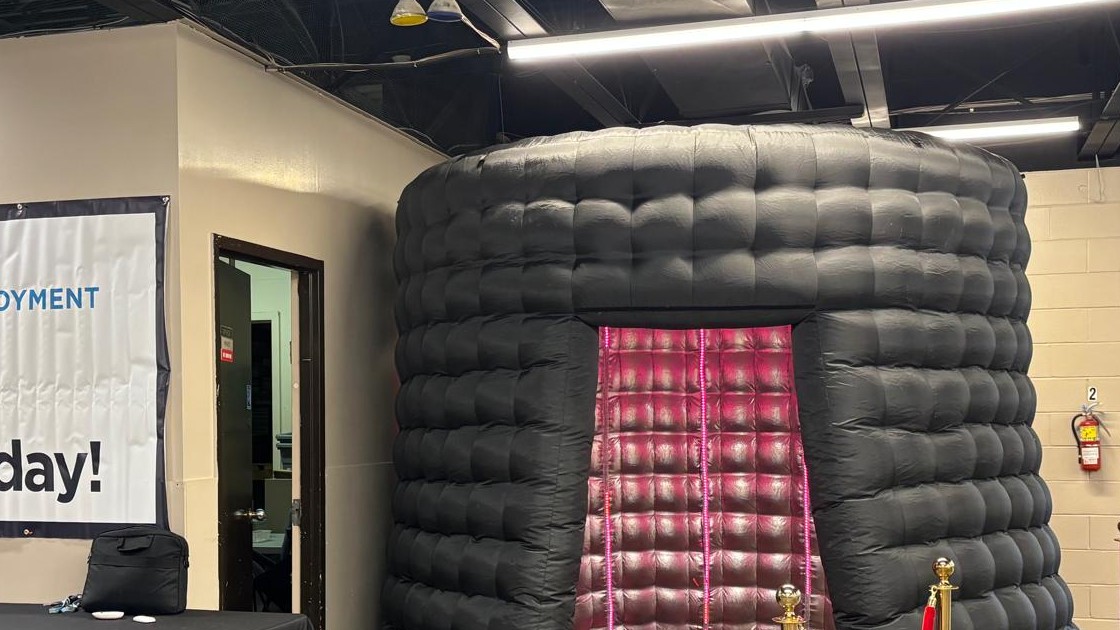 A black inflatable photo booth enclosure with a pink lit interior standing against a block wall