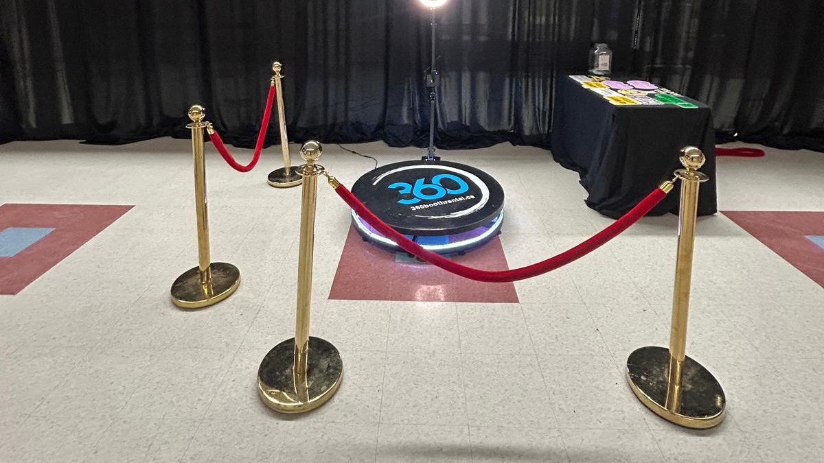A 360 booth on a lit platform with its ring light above, black pipe and drape behind it and a props table to one side