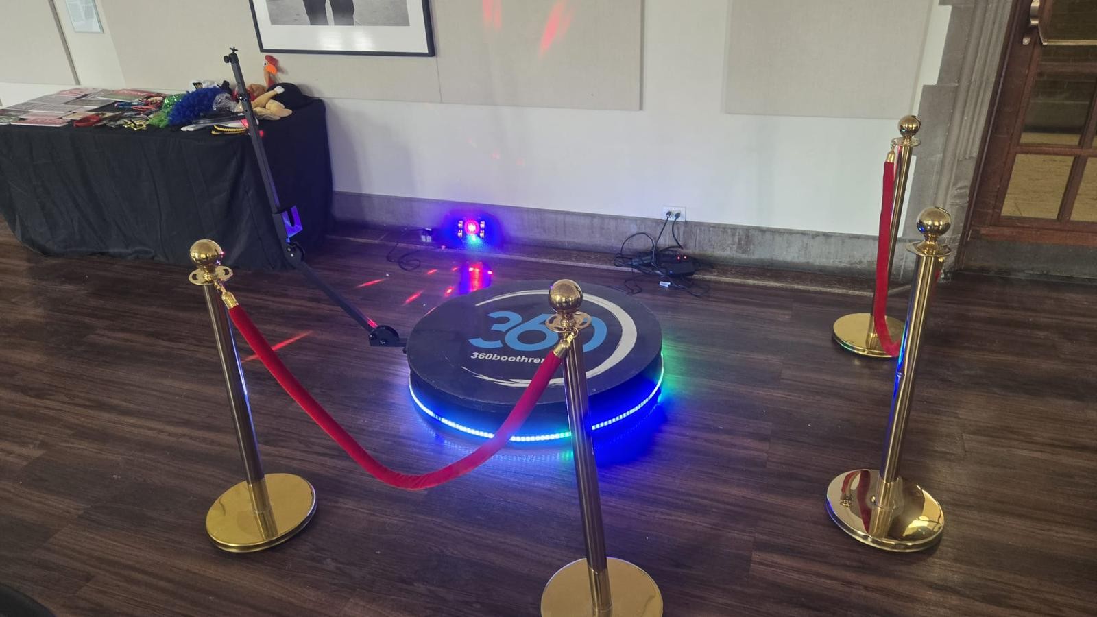 The 360 platform lit on a dark wood floor inside a rope line of four gold stanchions, with a props table behind
