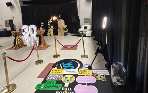 A red rope line leading to the lit 360 booth platform in a hall, with a props table in the foreground and lit marquee numbers across the room