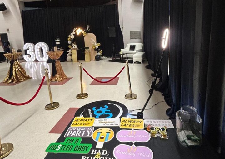 A red rope line leading to the lit 360 booth platform in a hall, with a props table in the foreground and lit marquee numbers across the room