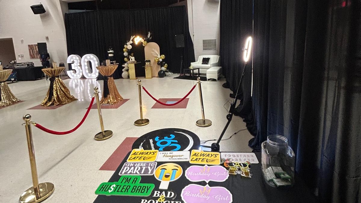 A red rope line leading to the lit 360 booth platform in a hall, with a props table in the foreground and lit marquee numbers across the room