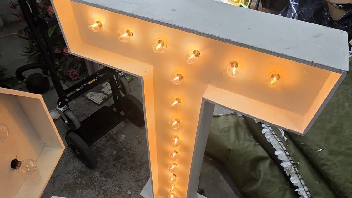 A single lit marquee letter lying in the workshop beside an unlit blank, a dolly and a dust sheet