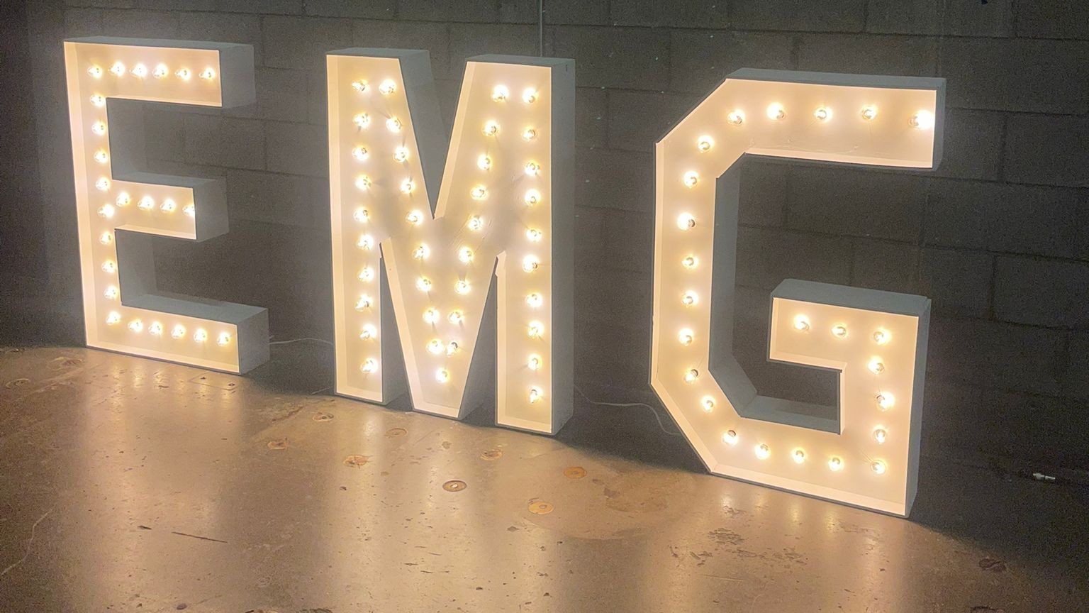 The letters EMG lit against a bare concrete block wall, reflecting on polished concrete floor