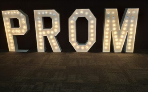 Four lit marquee letters spelling PROM standing against black drape on a dark carpet