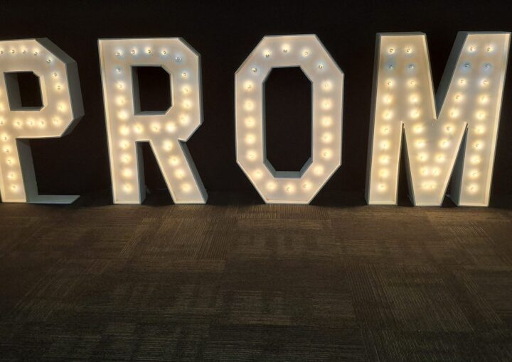 Four lit marquee letters spelling PROM standing against black drape on a dark carpet