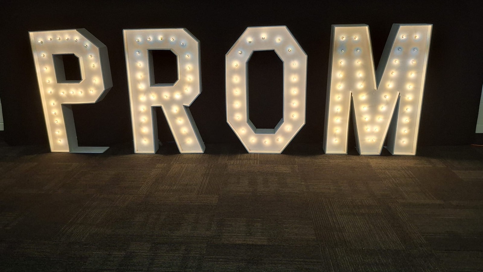Four lit marquee letters spelling PROM standing against black drape on a dark carpet