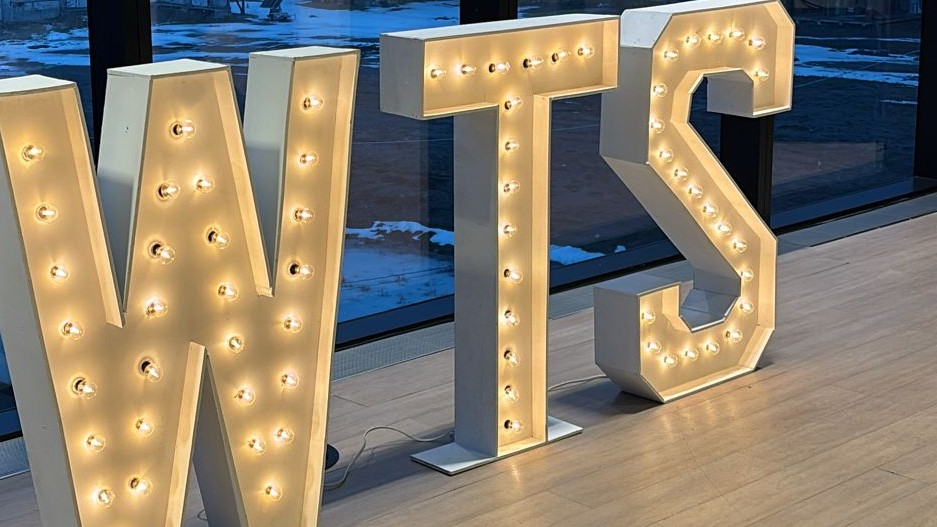 The letters WTS lit on their base plates on a pale wood floor in front of floor to ceiling glazing at dusk
